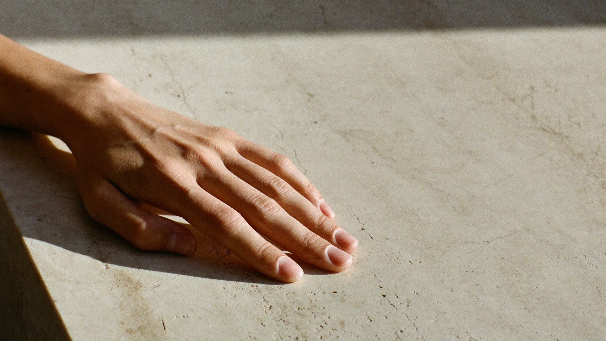 Elegante Hand mit natürlicher, ungeschminkter Maniküre auf hellem Travertinstein, weiches Seitenlicht